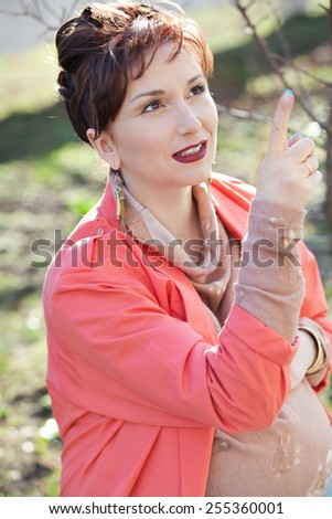 Beautiful young pregnant woman in a coral jacket stroking belly with hands and communicates with the unborn child at spring outdoor