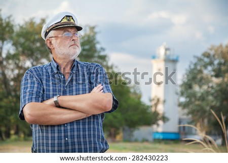 Elderly sea captain looks into the distance on sea beach against the backdrop of lighthouse