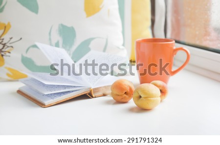 Three apricots on a window sill against the background of book, pillows and cup at the window on a rainy day. Set for a cozy summer reading.