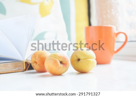 Three apricots on a window sill against the background of book, pillows and cup on the window. Set for a cozy summer reading.