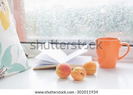 Three apricots on windowsill at the window with raindrops next to the book, pillow and cup on a rainy day. Set for a cozy summer reading.