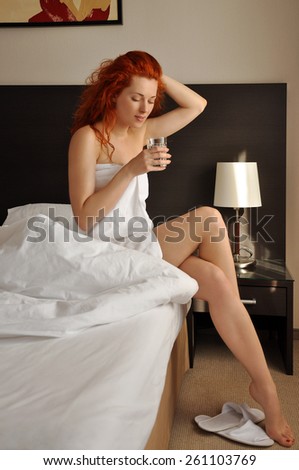 Red-haired girl sitting on the white bed with a glass of water