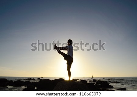 Asian man acts yoga on the beach, standing on one leg, silhouetted against sunlight.