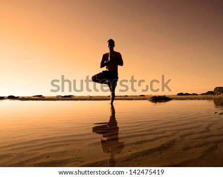 Asian man playing yoga on the beach, standing on one's leg, turn one side to the sunlight and other side in the dark. The scene sets on beautiful beach in Hua Hin, Thailand.