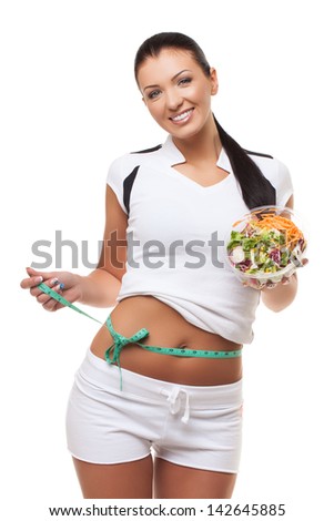Beautiful sporty healthy woman standing with mixed salad and measuring waist with centimetre tape over white background