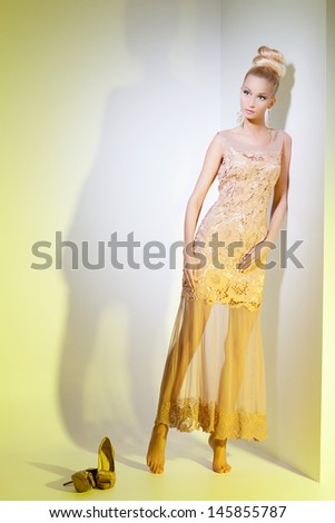 Portrait of beautiful blond girl in golden dress looking like doll leaning on wall over yellow background