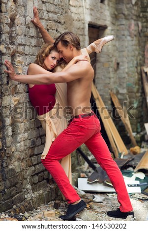 Dancer holding ballerina with her leg up against brick wall in trashed street