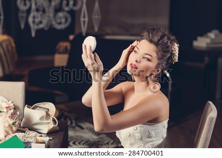 Beautiful young bride wearing fashion gown sitting on chair and looking into hand mirror