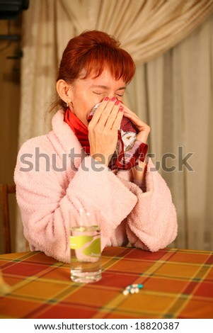 A sneezing woman sitting at the table with pills and a glass of water