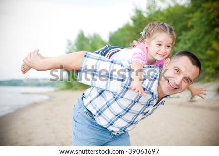 Middle-aged caucasian man playing with a little girl on the river bank. Man stands astoop ans holds his granddaughter on his back as she is \