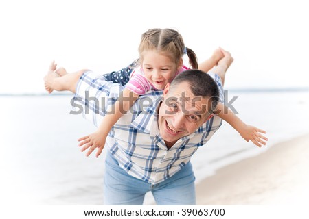 Middle-aged caucasian man playing with a little girl on the river bank. Man stands astoop ans holds his granddaughter on his back as she is \