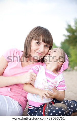 Portrait of happy young mother and daughter sitting on the sandy river bank one cool summer day. Mother embracing daughter. Girl is likely saying something amusing to mom.
