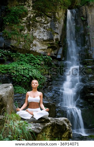 Beautiful young woman meditating in lotus position while doing yoga in the nature near waterfall