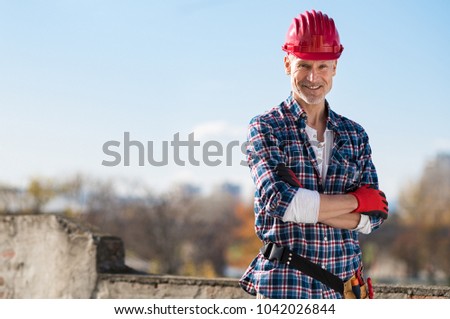 Smiling craftsman with arms folded looking at camera. Happy construction worker with tool kit on waist and work gloves standing on rooftop. Portrait of satisfied bricklayer with copy space.