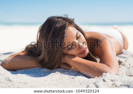 Relaxed woman lying down on sand during summer vacation. Beautiful girl lying down under the sun tanning in a tropical beach. Positive and serene young woman sunbathing at seaside with closed eyes.