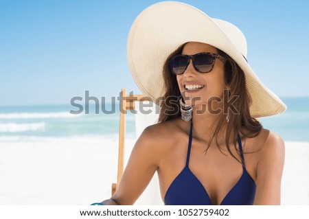 Young woman in blue bikini sitting on deck chair wearing white straw hat. Happy girl enjoying summer vacation at beach. Portrait of beautiful latin woman relaxing at beach with sunglasses.