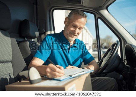 Happy delivery man with cardboard box checking list in van. Smiling courier checking list on clipboard. Deliveryman sitting in van and checking the delivery list.