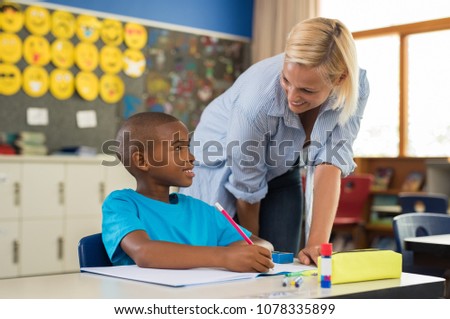 Teacher helping young african boy with homework. Happy teacher helping her student at elementary school. Portrait of woman teaching to schoolchild in classroom.