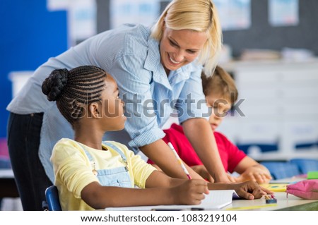 Happy teacher helping her students at elementary school. Teacher help schoolgirl and schoolboy writing test in classroom. Blonde teacher woman helping pupils studying at desk in classroom.