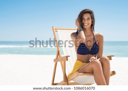 Happy smiling woman sitting on deck chair at sea. Young woman sunbathing in blue bikini while sitting with copy space. Laughing girl enjoying the sun at beach while looking at camera.