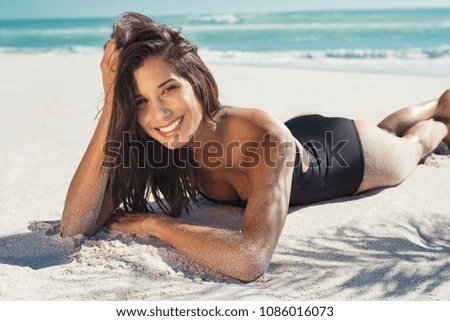 Happy young woman lying on white sand and looking at camera. Portrait of beautiful woman lying on front at tropical beach. Brunette girl in black swimsuit relaxing at sea under the palm trees.