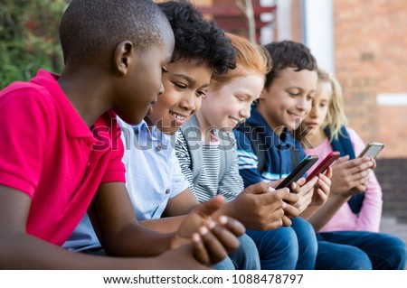 Pupils using mobile phone at the elementary school during recreation time. Group of multiethnic children sitting in a row and typing a message on smartphone. Young boys and girls with cellphone.