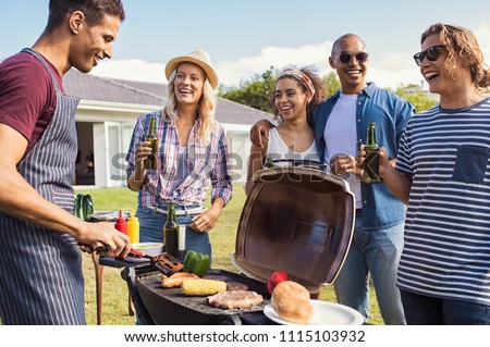 Multiethnic friends at barbecue party in the garden of house. Young man cooking meat on barbeque during summer holiday. Girls and guys having fun and drinking beers around the bbq.