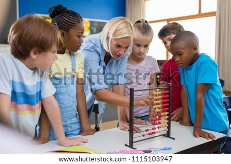 Teacher teaching children math on abacus. Mature mathematics teacher helping schoolchildren use wooden abacus. Group of multiethnic kids understanding maths in classroom.