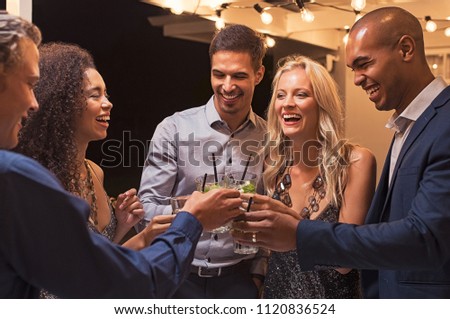Group of young multiethnic friends enjoying evening and drinking cocktails. Happy men and women raising a toast with mojito under the light bulb wire. Girls and guys having fun together at party.