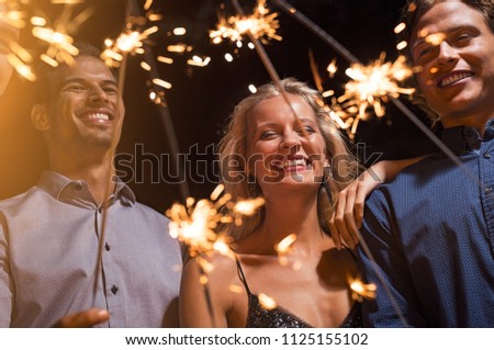Happy beautiful woman and men celebrating festival with bengal lights. Cheerful multiethnic group of friends enjoying with sparkling sticks. Smiling girl and guys celebrate new year’s eve.