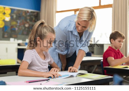 Happy teacher helping young girl with study in classroom. Teacher teaching to smiling schoolgirl at elementary school. Diligent child sitting at school desk with tutor.