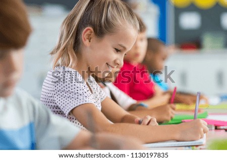 Smiling scholar girl sitting with other children in classroom and writing on textbook. Happy student doing homework at elementary school. Young schoolgirl feeling confident while writing on notebook.