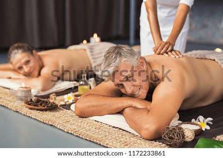 Mature couple in spa salon getting body massage and looking at camera. Senior man and beautiful woman relaxing at wellness center during beauty treatment. Mature couple lying on front at luxury spa.