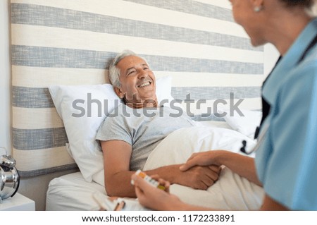 Cheerful senior man lying on bed receiving health care at home. Nurse advising old man on medication at home. Caregiver nurse helping elderly man taking medicine on the bed
