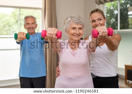 Happy senior couple exercising with dumbbells. Mature personal trainer helping elderly woman exercise at home. Old man and woman doing stretching exercise with physiotherapist.