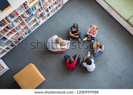 Teacher reading fairy tales to children sitting in a circle at library. Top view of librarian sitting with five multiethnic children on floor. Teacher reading book to girls and young boys at school.