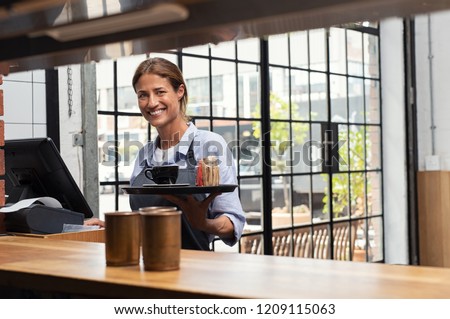 Smiling waitress serving cup of coffee in restaurant. Happy woman holding tray with cappuccino at the coffee shop and preparing bill while looking at camera. Latin woman making cheque for customer.
