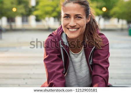 Portrait of smiling woman sitting on floor of city street after running. Healthy mature runner resting after workout exercise and looking at camera. Active sporty woman enjoying outdoors in autumn.