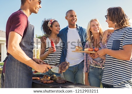 Young multiethnic friends having fun grilling meat enjoying bbq party. Group of happy guys and girls cooking and eating at barbecue dinner outdoor. Men and women standing around grill and chatting.