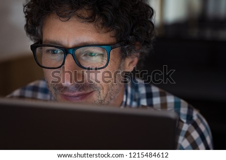 Mature man working on computer at night in dark office. Smiling business man wearing eyeglasses sitting at night working on laptop. Businessman wearing spectacles using digital tablet late at night.
