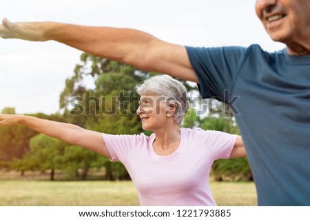 Senior woman stretching arms at park with her husband. Happy aged couple doing yoga exercise outdoor on a bright morning. Smiling elderly woman doing breath exercise with outstretched arms.