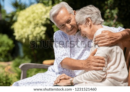 Senior man in medical patient cloth hugging wife outdoor bench. Old woman is consoled by her husband suffering from a terminal illness. Wife visit her husband at the hospital after the operation.