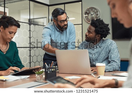 Latin mature manager discussing with his colleague online data in front of laptop. Serious businessman training employees in job. Creative multiethnic team working together with boss in meeting room.