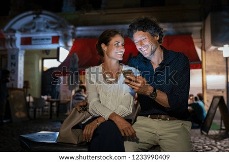 Man showing something to mature girlfriend in his smartphone outside the restaurant. Couple looking screen of smart phone. Happy woman and man having fun watching video on cellphone at night.
