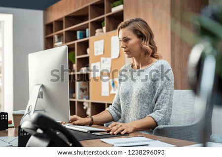 Mature businesswoman working on desktop computer at modern office. Concentrated freelancer finishing project sitting in coworking space using desktop computer. Creative business woman sending email.