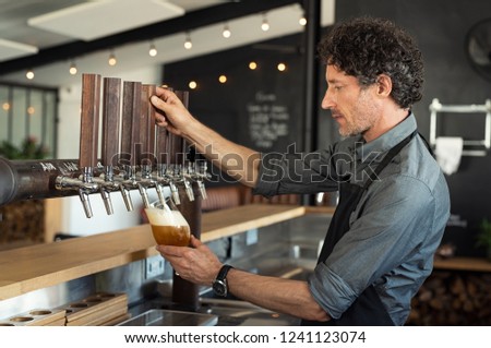 Mature bartender pouring beer in glass. Man pouring from tap fresh beer into the glass at pub. Side view of barman filling a glass with draught beer while standing at bar counter in restaurant.