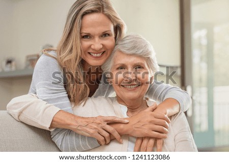 Cheerful mature woman embracing senior mother at home and looking at camera. Portrait of elderly mother and middle aged daughter smiling together. Happy daughter embracing from behind elderly mom.