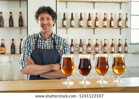 Smiling bartender wearing apron standing behind counter with beer bottles on shelves and craft beer in glasses at counter. Portrait of cheerful barman standing with different types of draught beer.