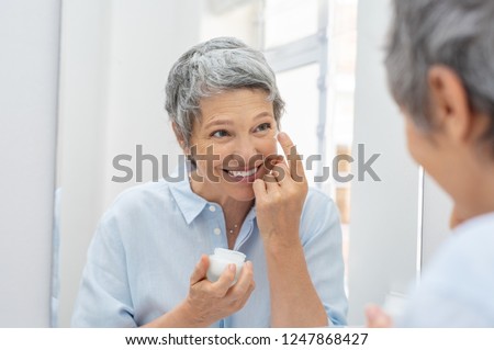 Happy mature woman applying face lotion while looking herself in the bathroom mirror. Senior woman applying anti aging moisturizer on her face. Smiling lady holding little jar of skin cream.