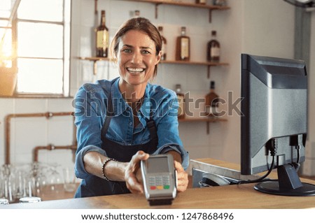 Waitress standing at cash counter holding an electronic card payment machine. Smiling mature woman holding terminal machine and looking at camera.